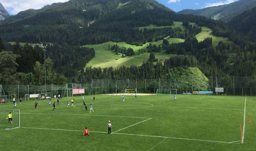 Partida de futebol em campo verde com paisagem de montanha ao fundo