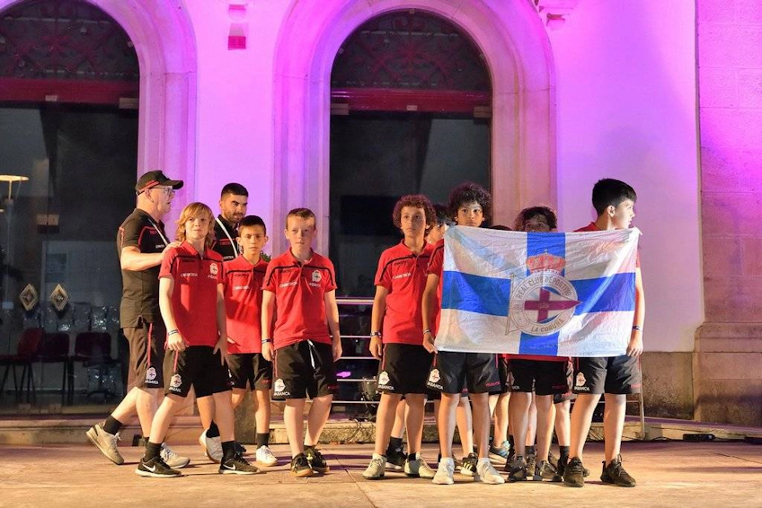 Группа юных футболистов в красной форме с тренером, держащих флаг, на ночном спортивном мероприятии.