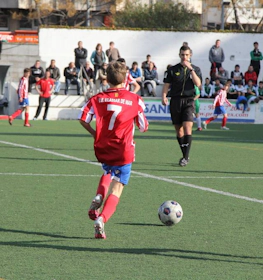 Young player number 7 in red controlling the ball with a referee and spectators in the background.
