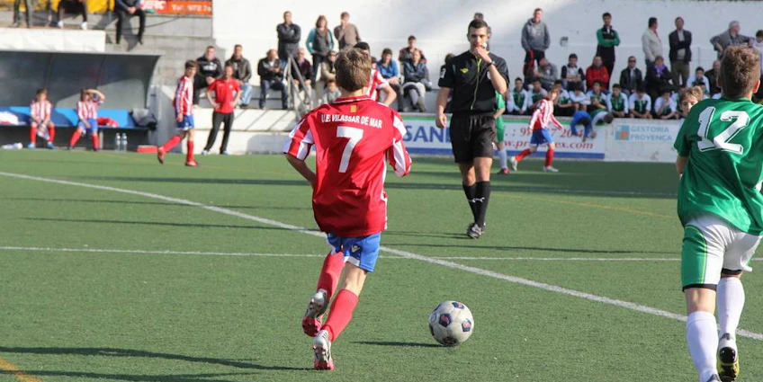 Молодой игрок номер 7 в красной форме ведет мяч на турнире Bahia de Roses Cup.