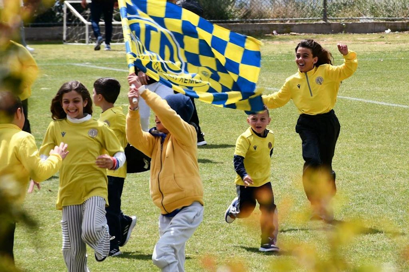 Дети в желтых футболках радостно бегут по футбольному полю с флагами