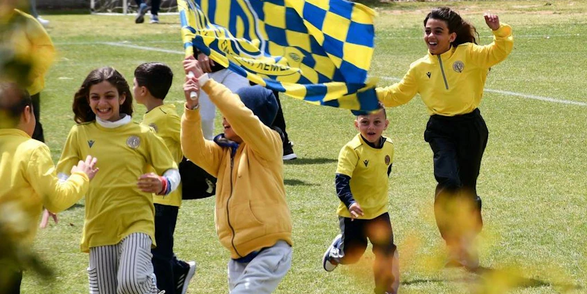 Sarı formalı neşeli çocuklar bayraklarla futbol sahasında koşuyor