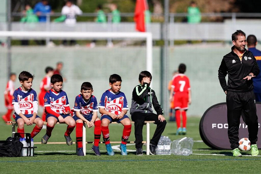 Genç futbolcular ve antrenör, FIT 24 Promises Edition turnuvası sırasında saha kenarında yedek kulübesinde oturuyor
