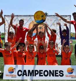 Genç futbol takımı Valencia Beach Turnuvası'nda antrenörler ve 'Campeones' pankartı ile zafer kutluyor.