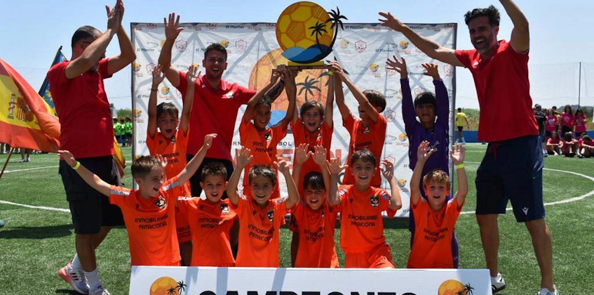 Юные футболисты празднуют победу на турнире Valencia Beach с тренерами и баннером 'Campeones'.