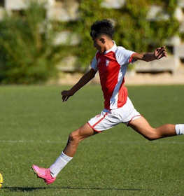 Jogador jovem em uniforme branco e vermelho executando um chute poderoso durante um jogo de futebol em um dia ensolarado.